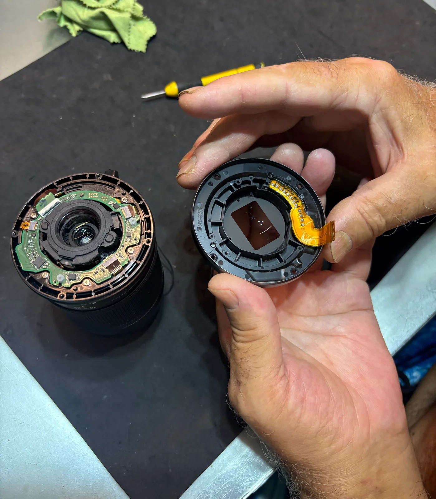 Technician holding part of a disassembled camera lens beside the lens barrel on a workbench.