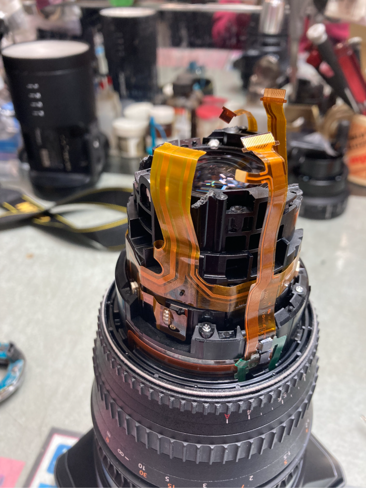 Disassembled camera lens with orange ribbon cables on the AL-ART repair bench.
